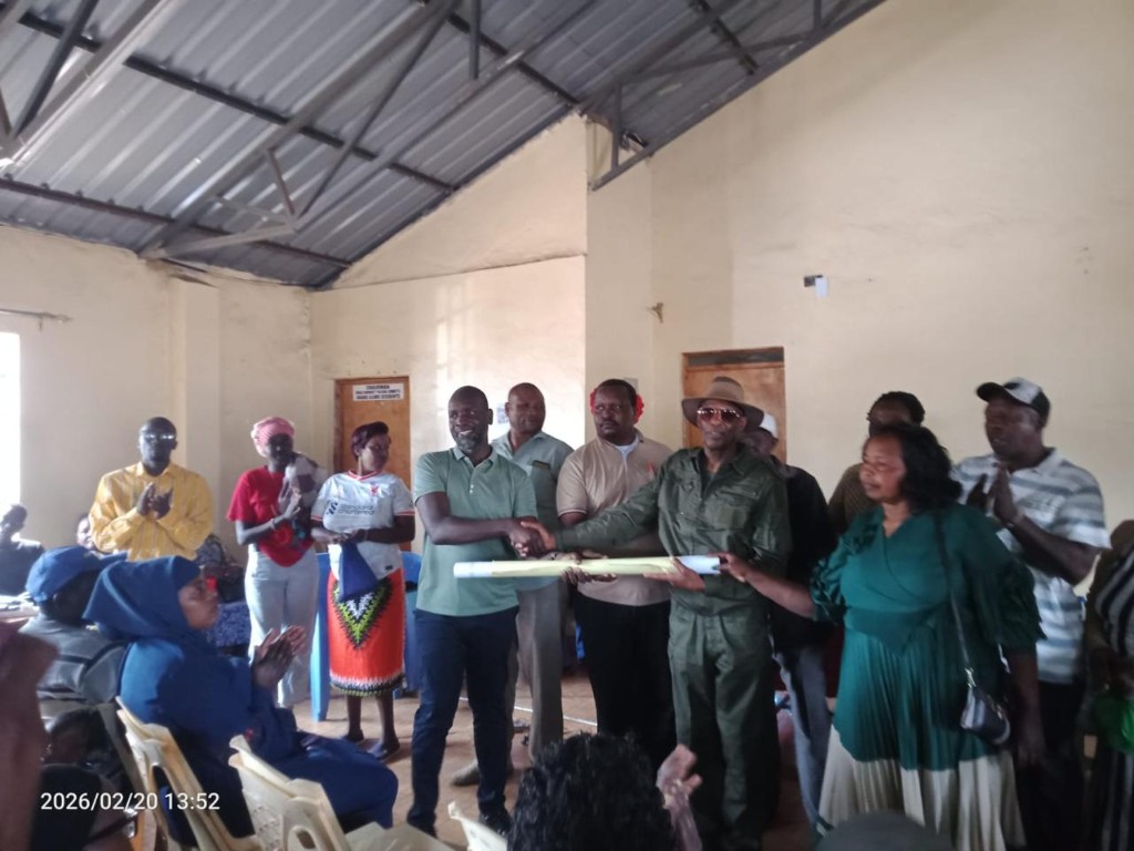 Formal handover of rolled programme documentation between KISRP representatives and community leaders during an indoor ceremony